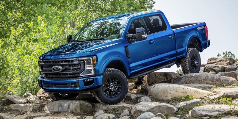 A blue Ford F-250 Super Duty pickup truck with large tires climbs over rocks in a wooded area.