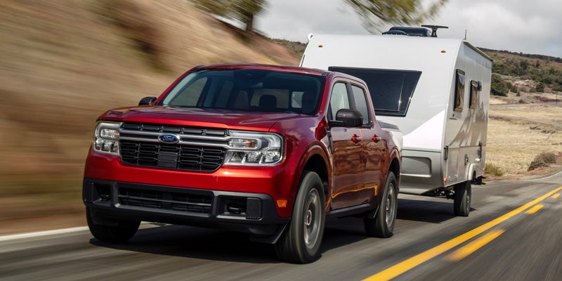A red Ford Maverick pickup truck tows a white camper trailer on a winding road with blurred motion.