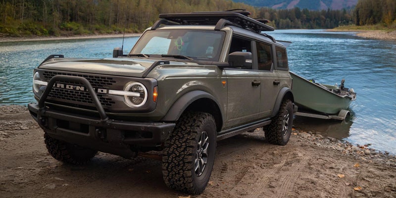 Dark green Ford Bronco with a roof rack on a dirt riverbank, towing a small boat into the blue river.