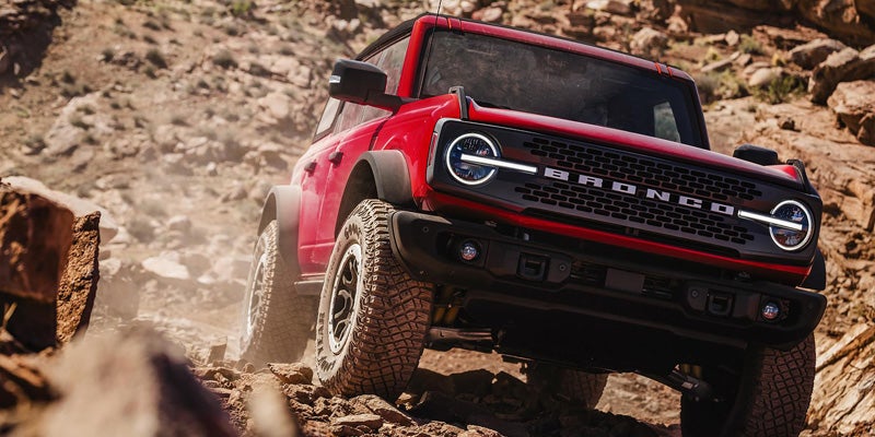 A red Ford Bronco driving uphill on a rocky, dusty off-road trail with sunlight.