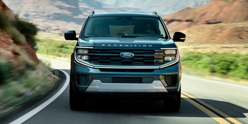 Front view of a dark blue Ford Expedition driving on a winding road with rocky hills and green vegetation.